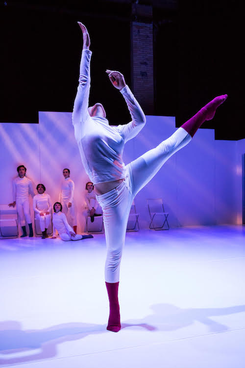 Dancers huddle together against a white wall while they look over to a dancer who extends their left leg high into the air